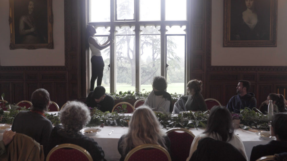 A performer stands of a windowsill inside Ashton Court Mansion. An audience seated at a long dining table watch.