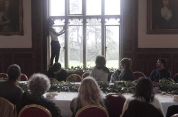 A performer stands of a windowsill inside Ashton Court Mansion. An audience seated at a long dining table watch.