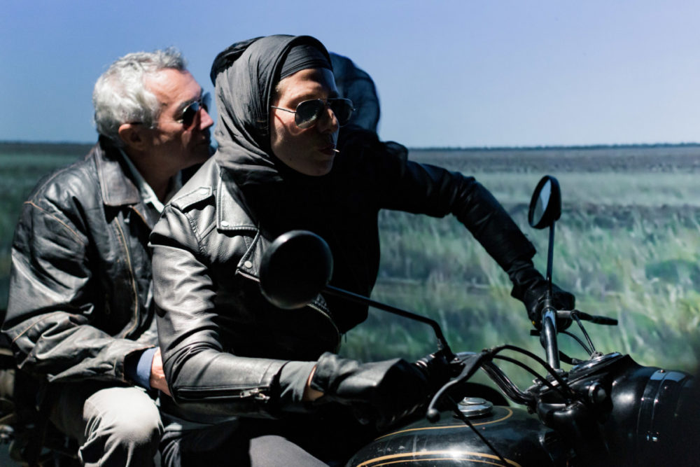A woman in a headscarf rides a motorbike, a man with grey hair sits on the back