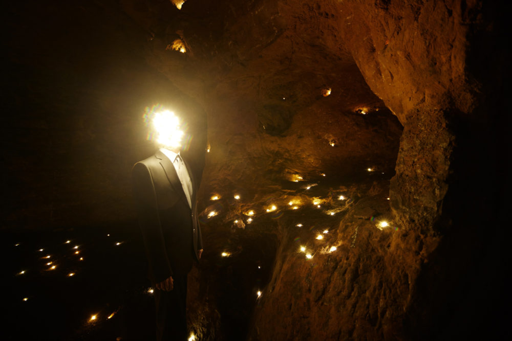 A man with a face made of lights stands in a cave which contains lots of tiny lights stands in Redcliffe Caves