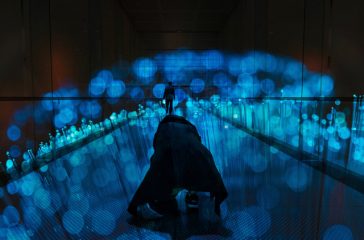 A photo of a figure kneeling in a dark space, on a dark reflective floor, lit by blue lights