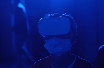 A close up photograph of a man wearing vr headset goggles and a face mask. The lighting is dark blue.